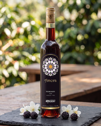 Botella de hidromiel marca Amora de Costa Rica Meadery sobre una mesa de madera al aire libre, decorada con zarzamoras frescas y flores blancas en la base.